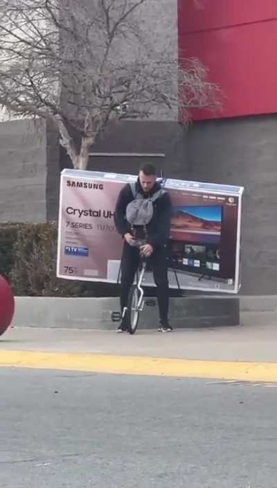 Dude rides off on a unicycle carrying a new 75" television on his back