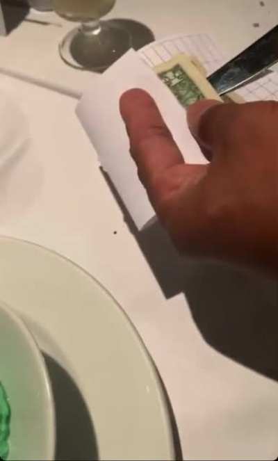 Waiter only cuts a hole in the paper and not the dollar but manages to put the knife through the dollar..