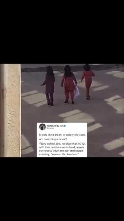 Young school girls, no older than 10-12 years old, with their headscarves in hand, march confidently down a street in Iran while chanting, “Women, Life, Freedom!”